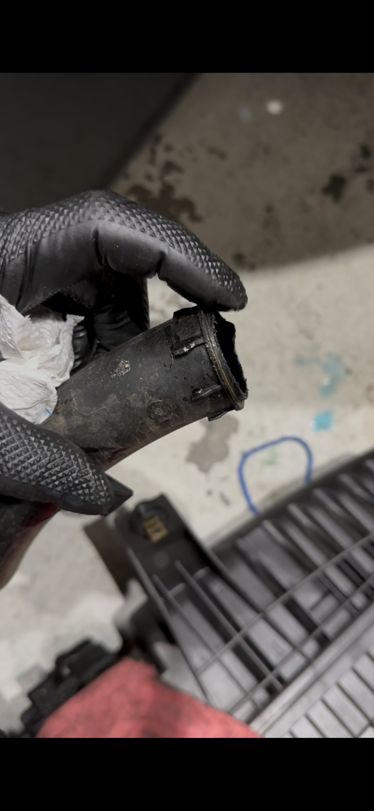 Gloved hand holding the removed plastic coolant crossover pipe, showing the open water-pump end with a ragged, eaten-away section of the sealing rim