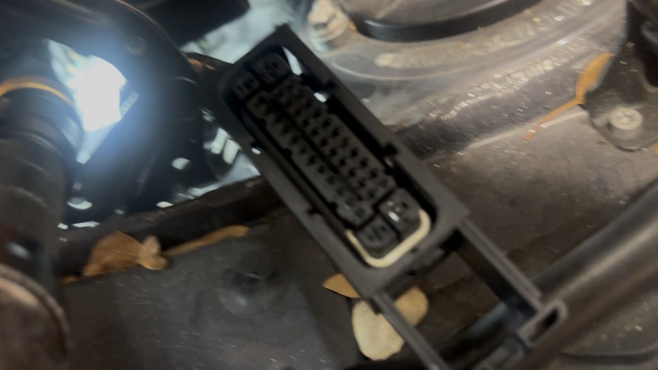 Multi-pin control unit connector held up in an engine bay, lit by a work light